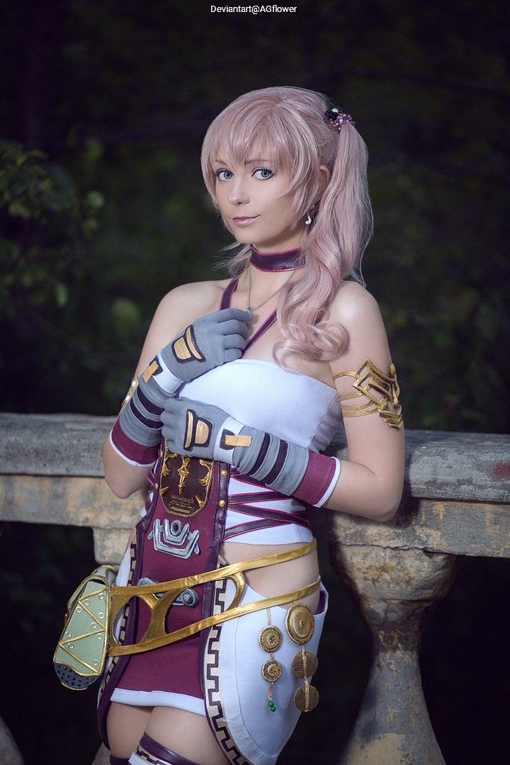 A person in a fantasy costume with long light pink hair stands by a stone railing in a natural setting, showcasing intricate attire with accessories.