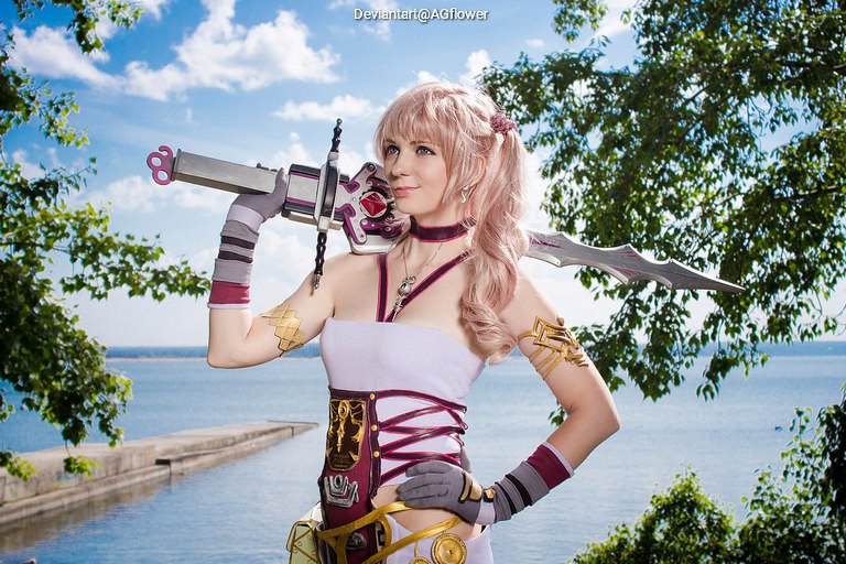 A woman in a fantasy-themed costume poses confidently by the water, holding a stylized sword. She has long, light-colored hair and wears accessories that complement her outfit against a sunny backdrop of trees and water.