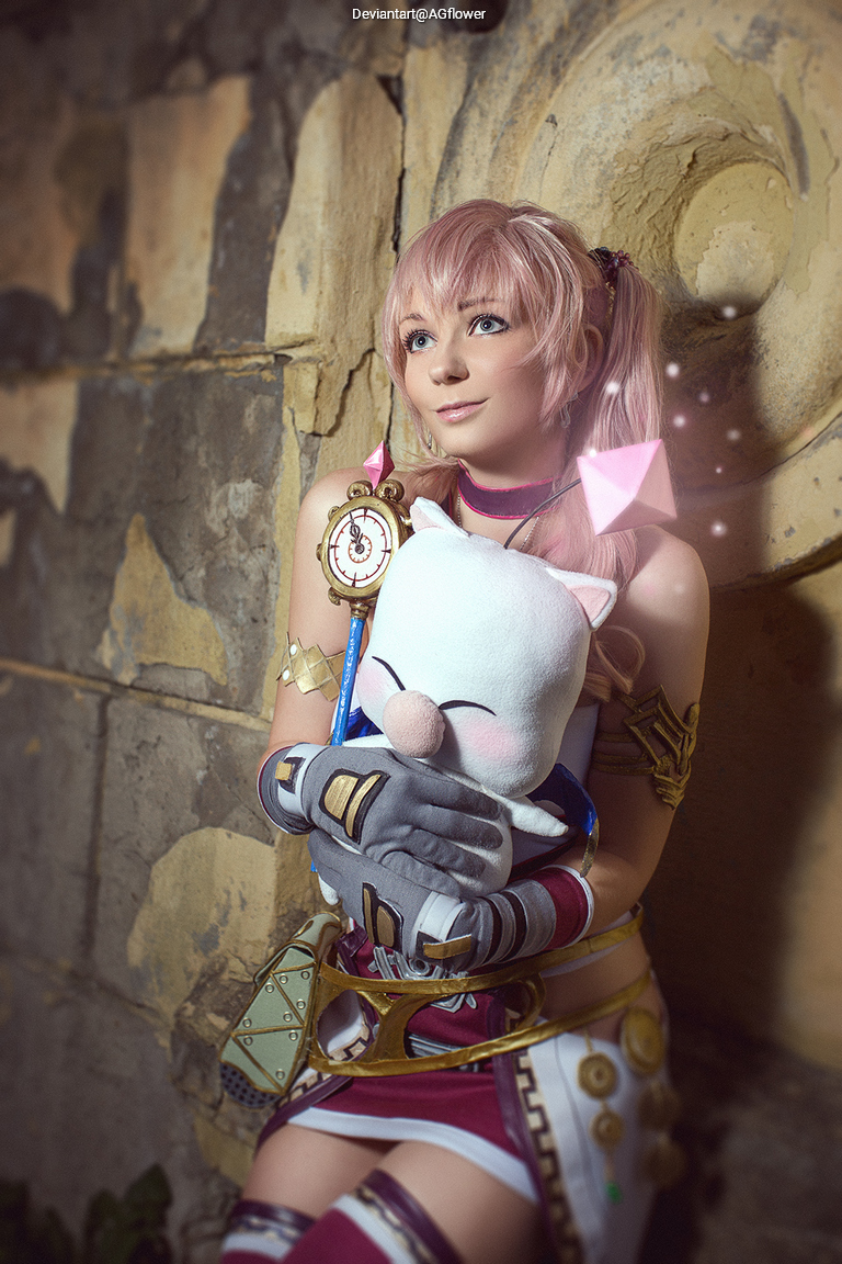 A person with pink hair dressed in a fantasy-style outfit holding a plush toy, posing against a textured stone wall.