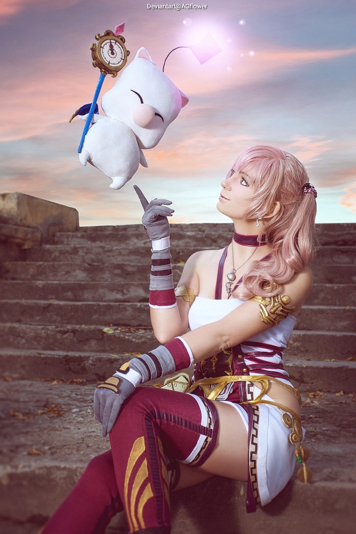 A character in costume resembling Serah Farron from a fantasy game sits on steps, playfully interacting with a small, fluffy creature resembling a Moogle, set against a colorful sunset backdrop.