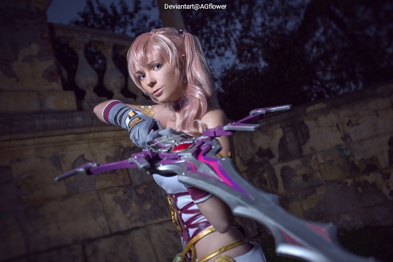 A cosplayer with pink hair, dressed in a fantasy-themed outfit, poses with a large, ornate weapon against a night backdrop.