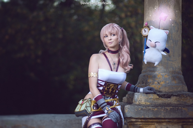 A woman in a fantasy outfit inspired by a video game character sits next to a stone pillar, with long pink hair and a white top embellished with intricate patterns. She holds a small, friendly creature resembling a Moogle from a popular game series.