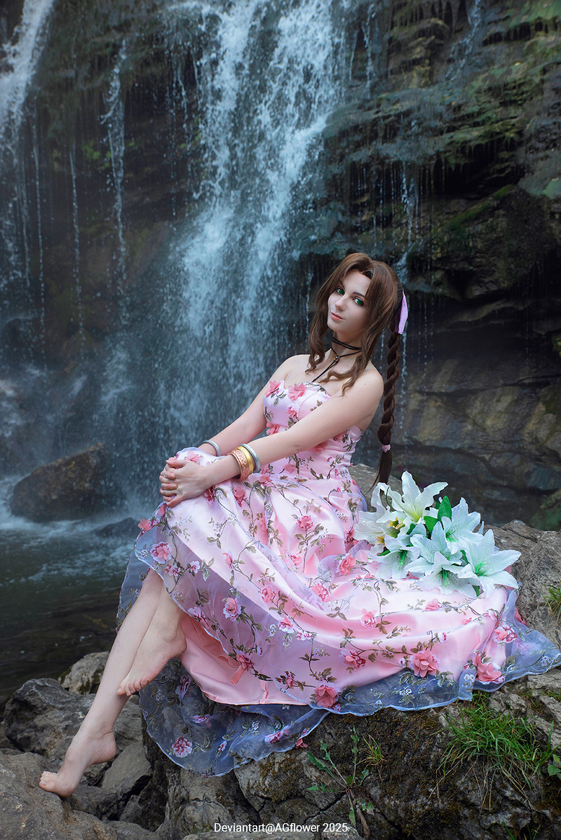 A young woman in a pink floral dress sitting on a rock near a waterfall, holding white flowers.