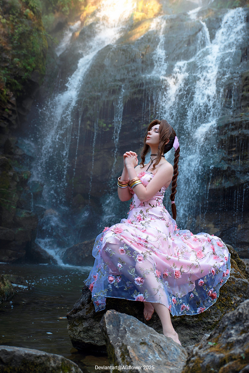A person in a floral dress sits on a rock near a waterfall, with hands clasped in prayer, surrounded by lush greenery.