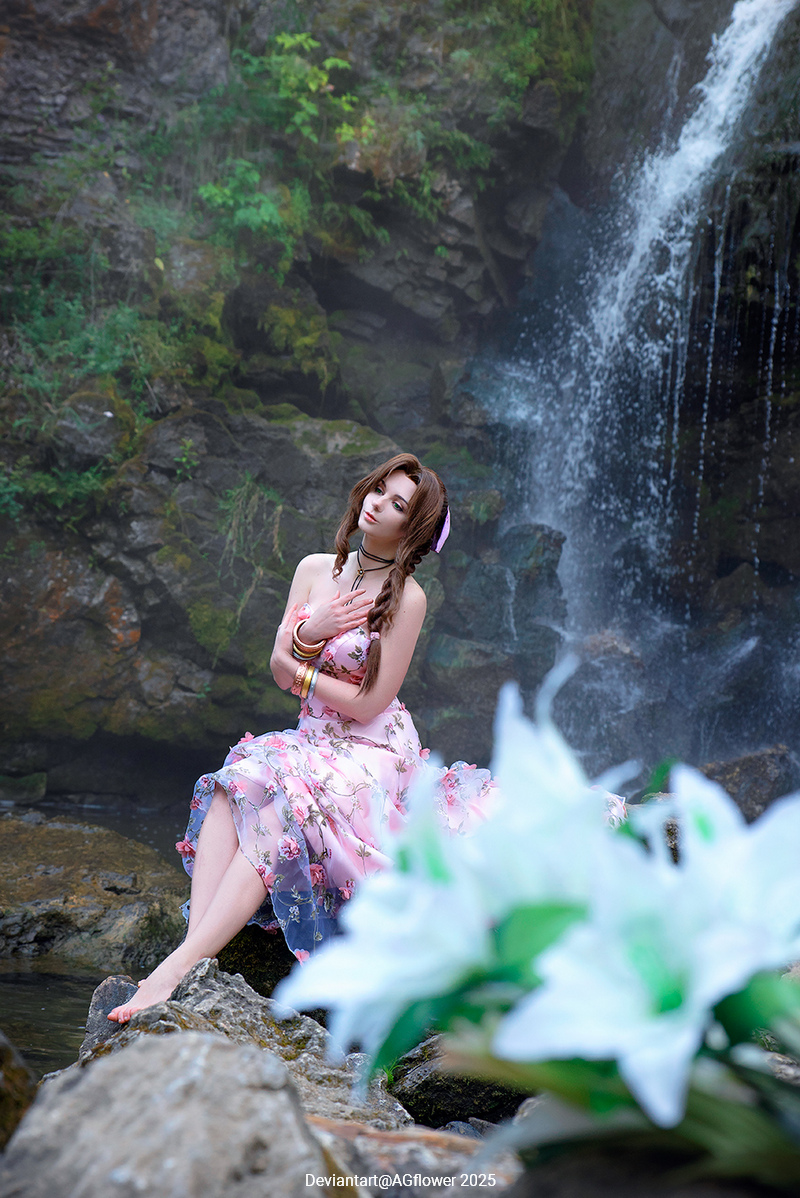 A person in a pink floral dress sits on a rock by a waterfall, surrounded by greenery and flowers.