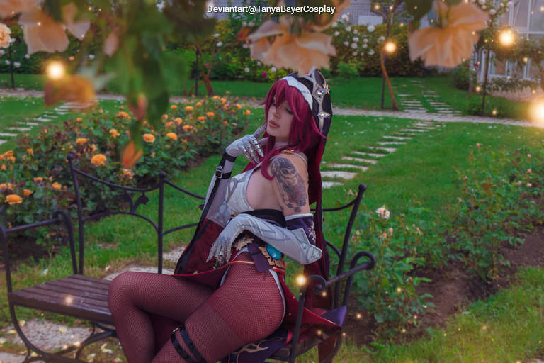 A person in cosplay sitting on a park bench surrounded by roses, wearing an elaborate costume with long red hair and a tattoo visible on their arm, under soft, ambient lighting.