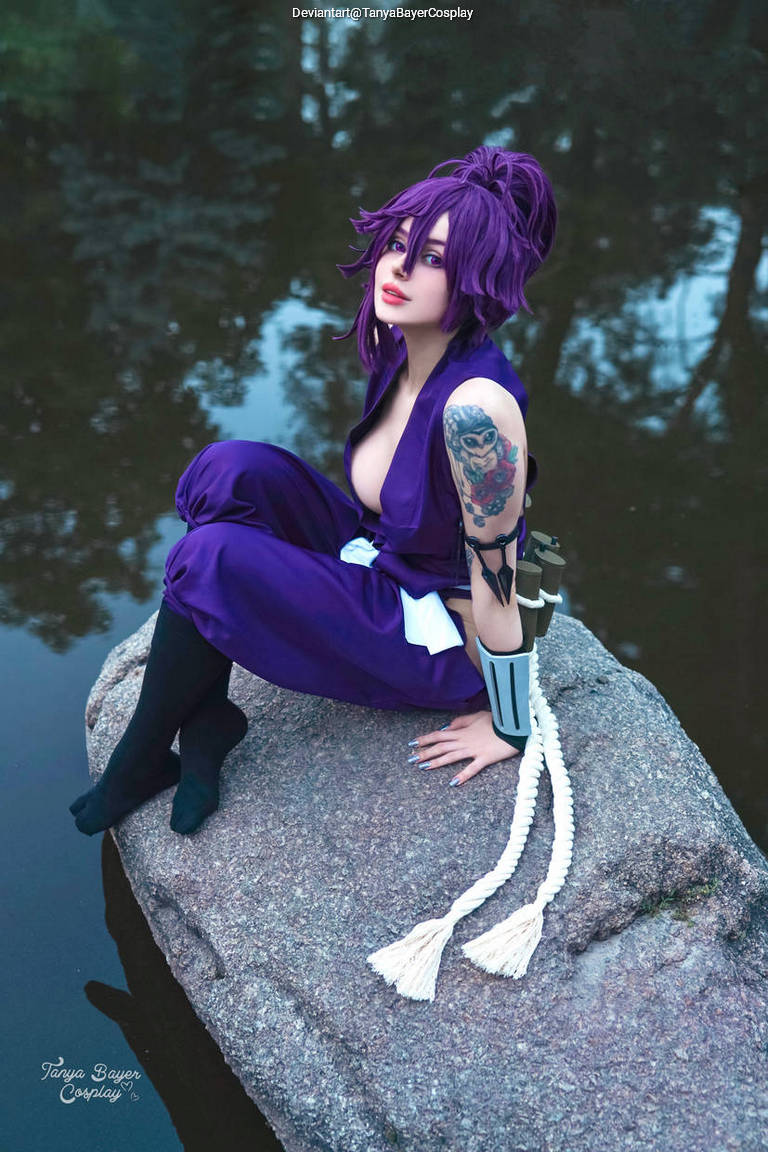 A person in a purple outfit with short purple hair poses on a rock by a calm body of water, showcasing a tattoo on their arm and wearing black stockings.