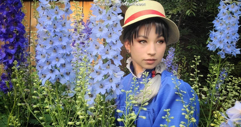 A person wearing a blue outfit and a straw hat stands among tall blue flowers in a garden setting.