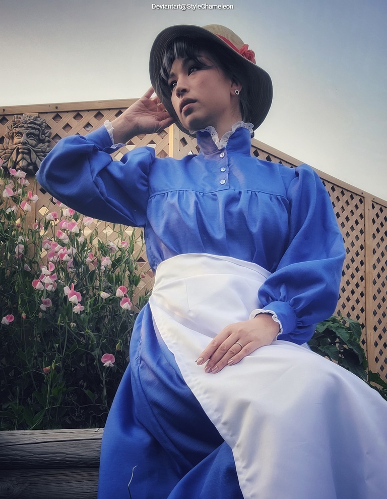 A woman in a blue vintage dress with an apron, wearing a straw hat, sitting and gazing thoughtfully in a garden setting with blooming flowers in the background.