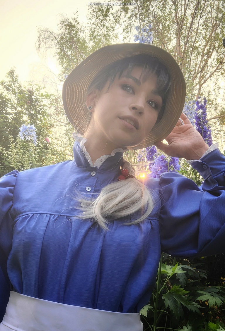 A person wearing a blue blouse and a straw hat, posing amid green plants and colorful flowers.