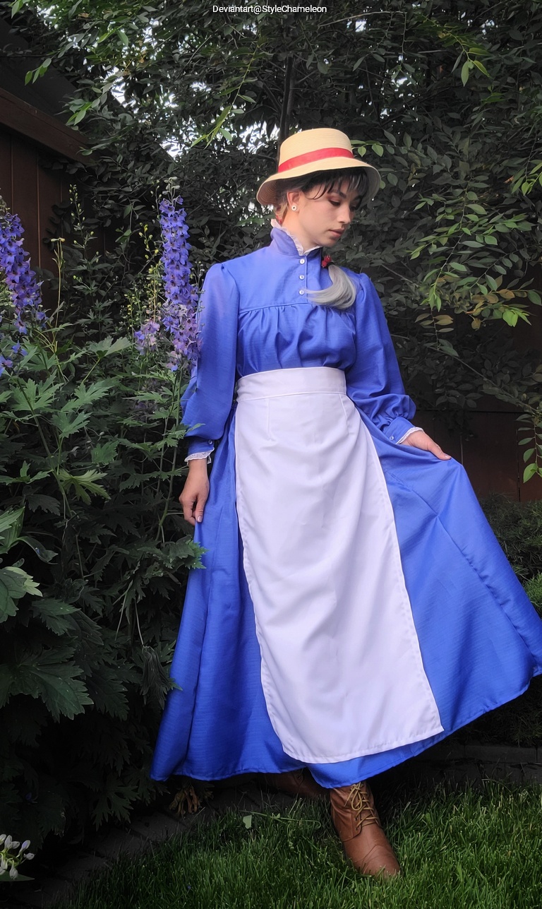 A person wearing a blue vintage-style dress with a white apron, standing in a garden with purple flowers and greenery, wearing a straw hat.