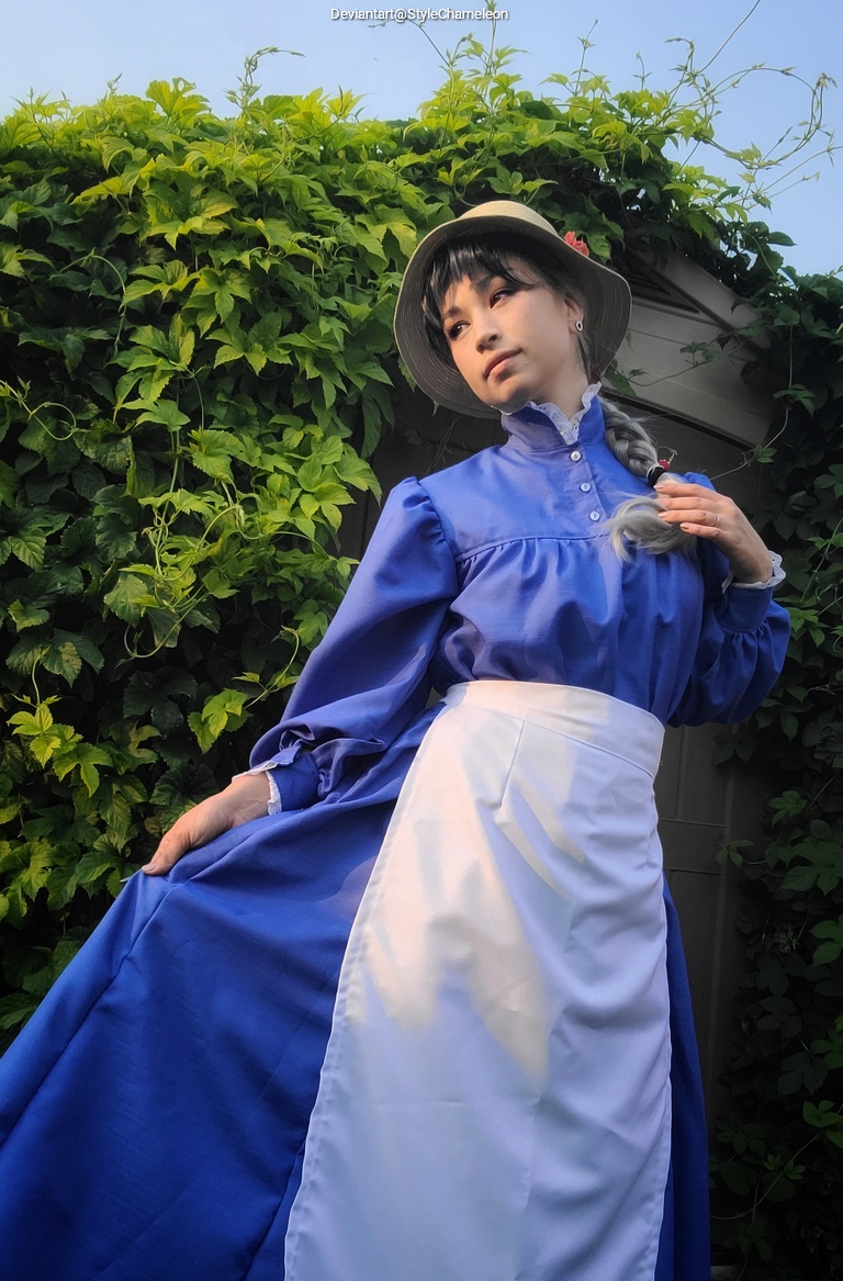 A person wearing a blue dress and a straw hat, posed in front of a lush green backdrop of leaves.