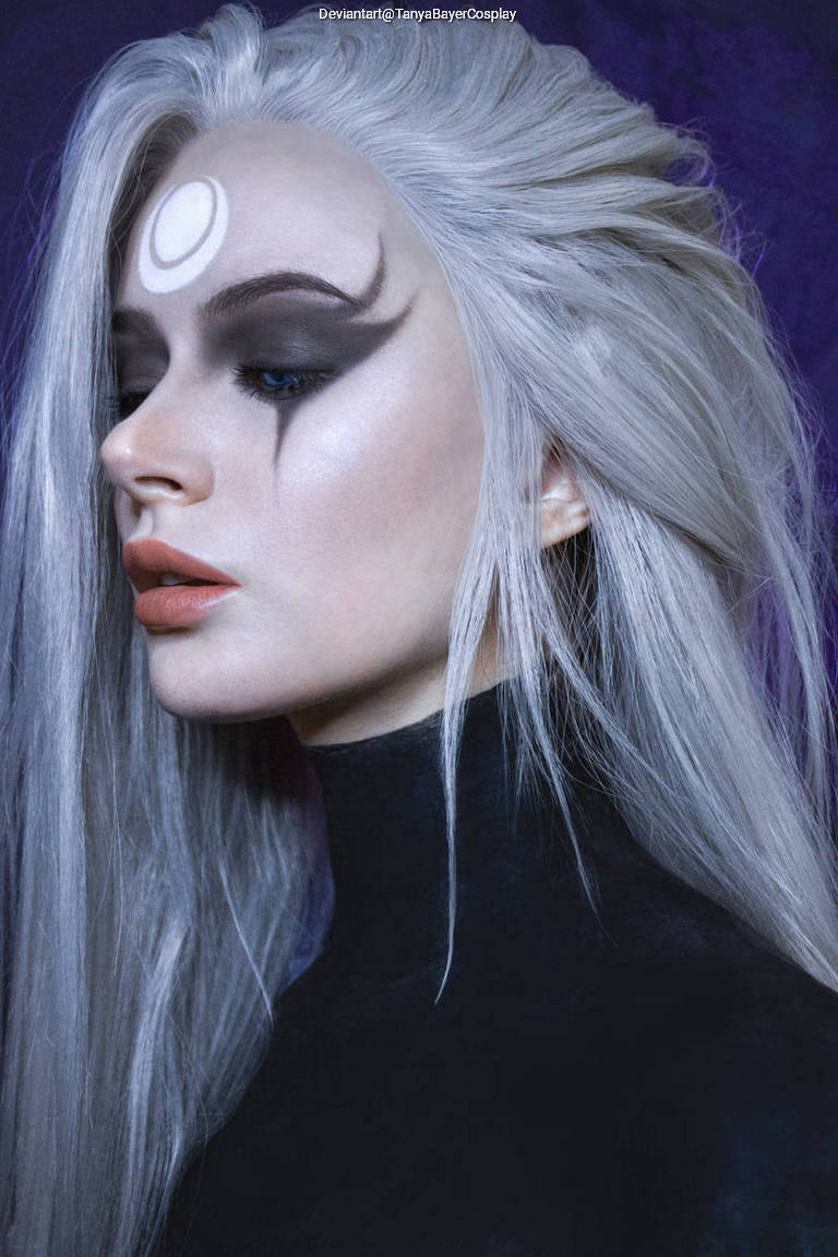 Profile view of a person with long, silver hair and dramatic makeup, featuring dark eyeshadow and a white crescent symbol on their forehead.