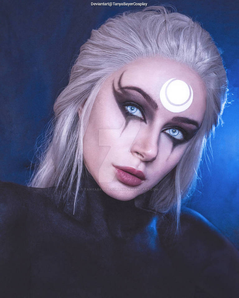 A close-up portrait of a person with long, light-colored hair, wearing dark makeup with dramatic eyeliner. The individual has striking blue eyes and a moon symbol on their forehead, conveying a mystical or fantasy theme.