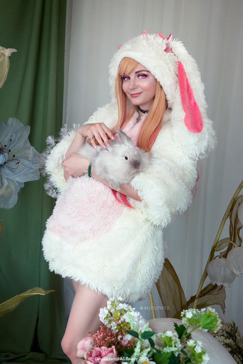 A person with long, light orange hair wearing a fluffy white coat with bunny ears, holding a small rabbit in their arms, surrounded by flowers and greenery.