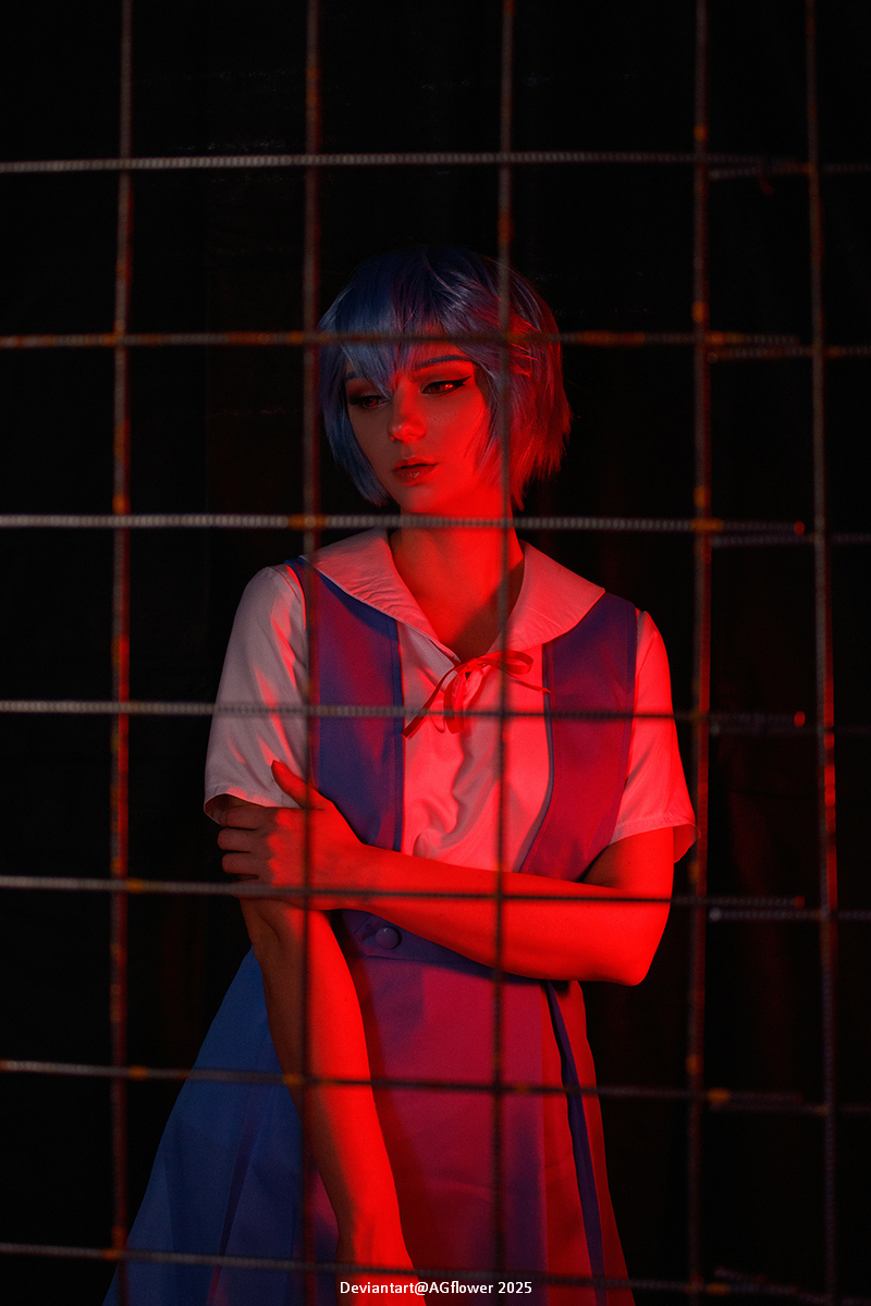 A person with short blue hair, wearing a light blue dress with a white collar, poses in front of a grid with red lighting.