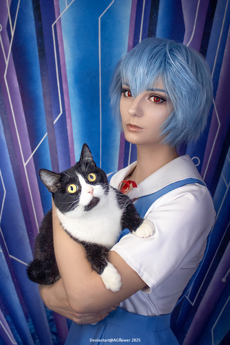 A person with short blue hair and red eyes holds a black and white cat, standing in front of a blue and geometric patterned backdrop.