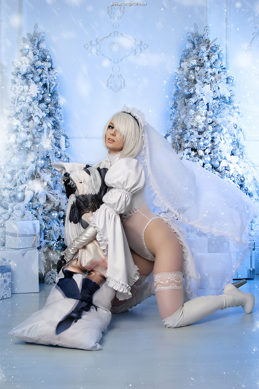 A person in a white costume with a veil is posing in a winter-themed setting with decorated evergreen trees and gift boxes.