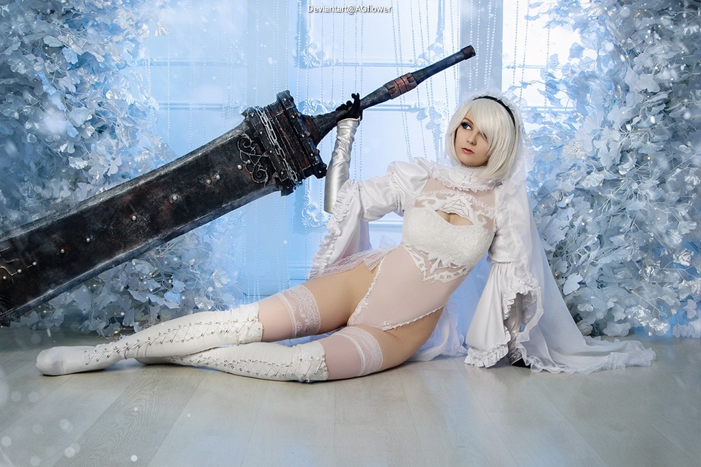 A person dressed in a white, intricately designed costume holds a large, ornate sword while sitting on the floor against a backdrop of frosty decorations.