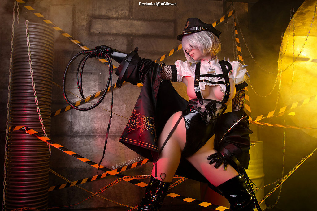 A model in an elaborate costume featuring a short, ruffled top with fishnet accents, black latex shorts, and thigh-high boots. She holds a whip and poses dramatically in a dimly lit industrial setting with caution tape and metal barrels in the background.