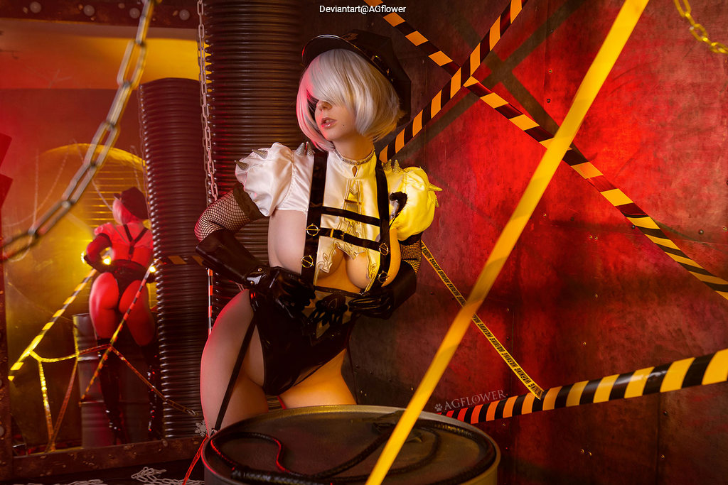 A person dressed in a provocative costume with pink hair, posing in an industrial setting with bright lighting and caution tape.