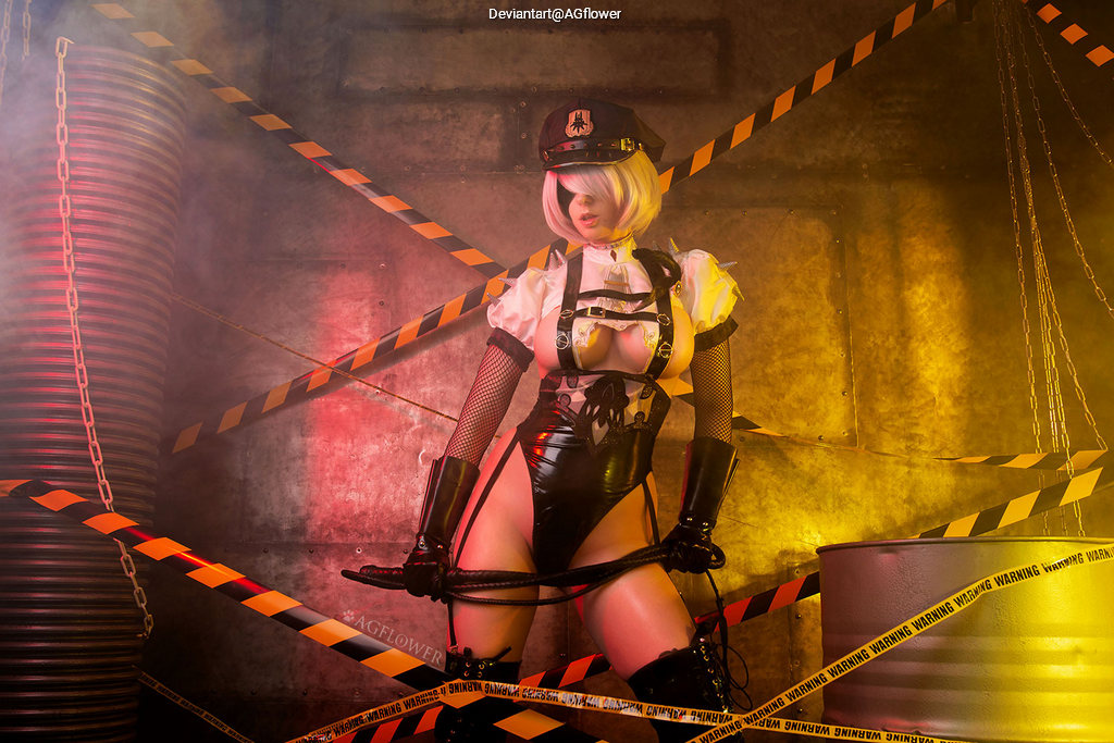 A character in a costume with a black hat and striking outfit, posed in a moody, industrial setting with warning tape and dramatic lighting.