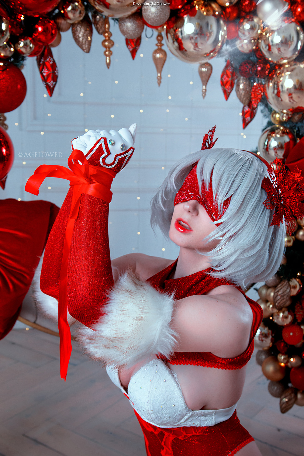 A person dressed in a festive red and white outfit, with a white wig, posing in front of a Christmas-themed backdrop adorned with ornaments and decorations.