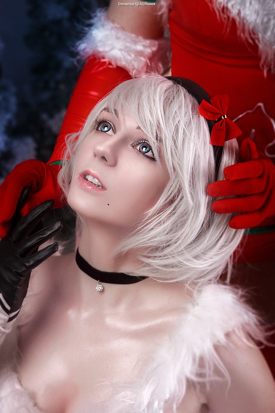 A portrait of a person with short white hair, wearing a festive costume with a fur trim, adorned with a black choker and a small decorative pendant. The person looks up with bright blue eyes, while another person in a red outfit holds their hair.