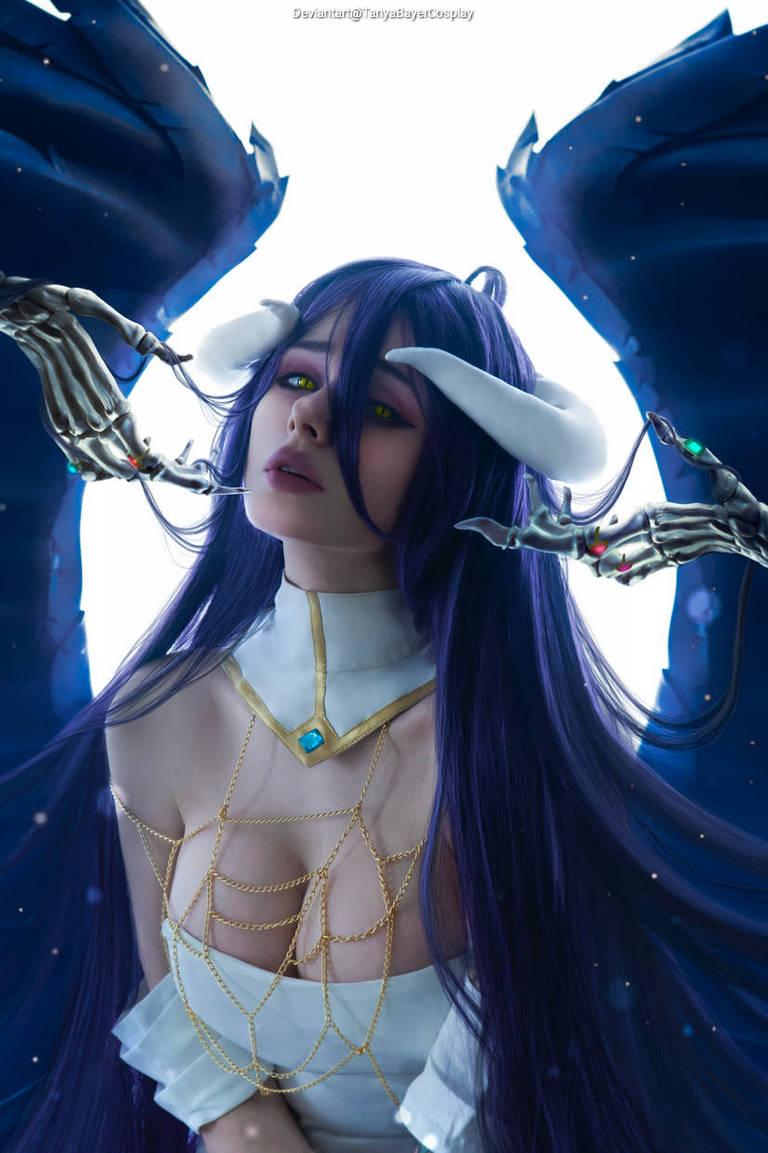 A female character with long purple hair and yellow eyes, dressed in an elegant outfit with gold accents, is surrounded by ethereal blue wings and skeletal hands reaching towards her face.