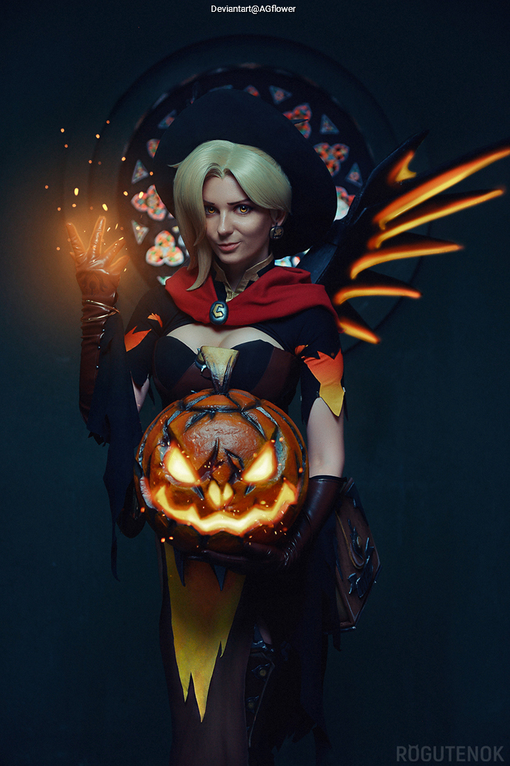 A woman dressed as a witch cosplaying with a glowing pumpkin, wearing a dark costume with colorful accents and a large hat, set against a dark backdrop with stained glass.
