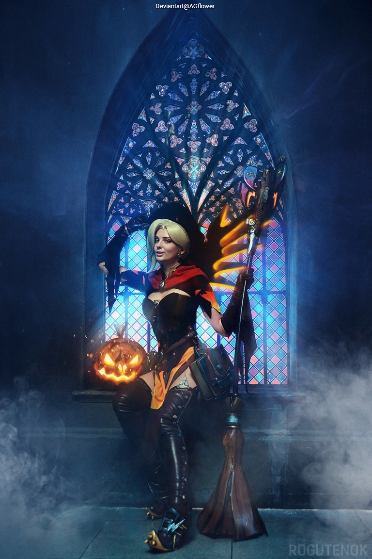A woman dressed as a witch holds a carved pumpkin while standing in front of a colorful stained glass window, surrounded by mist.