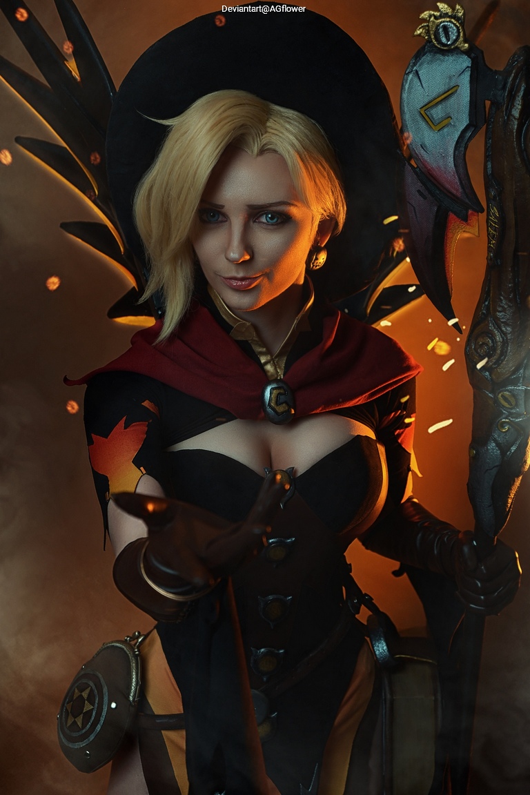 A female cosplayer dressed as a character with a large black hat and a red cloak, holding a weapon, standing against a dramatic lit background.