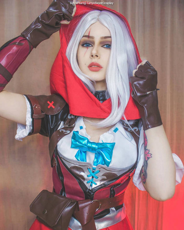 A person in a detailed costume featuring a red hood and leather accessories, posing with a dramatic expression. The outfit includes a white blouse and a blue bow, with visible tattoos on one arm.