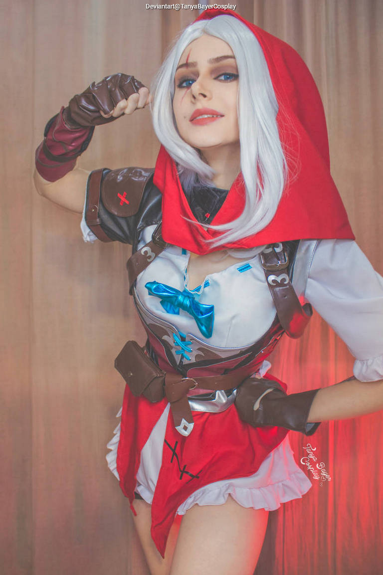 A woman in a costume with a red hood and a playful expression, showcasing intricate details on her outfit, including a blue bow and various accessories.