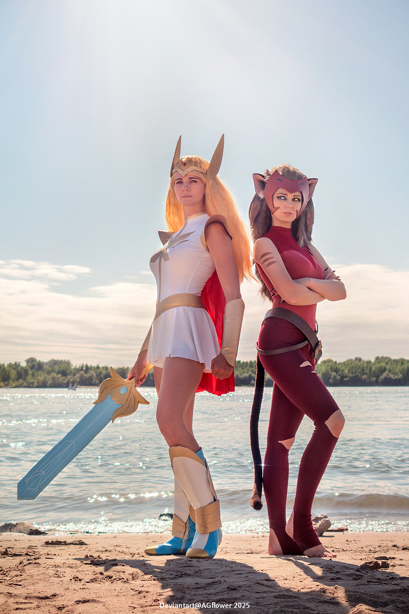 Two women in cosplay standing on a beach, one dressed as She-Ra with a sword and the other as Catra with cat-like features.
