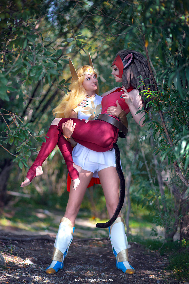 Two women in cosplay standing in a lush green setting, one dressed as She-Ra with blonde hair and armor, the other as Catra with dark hair and red attire, with Catra being held in She-Ra's arms.