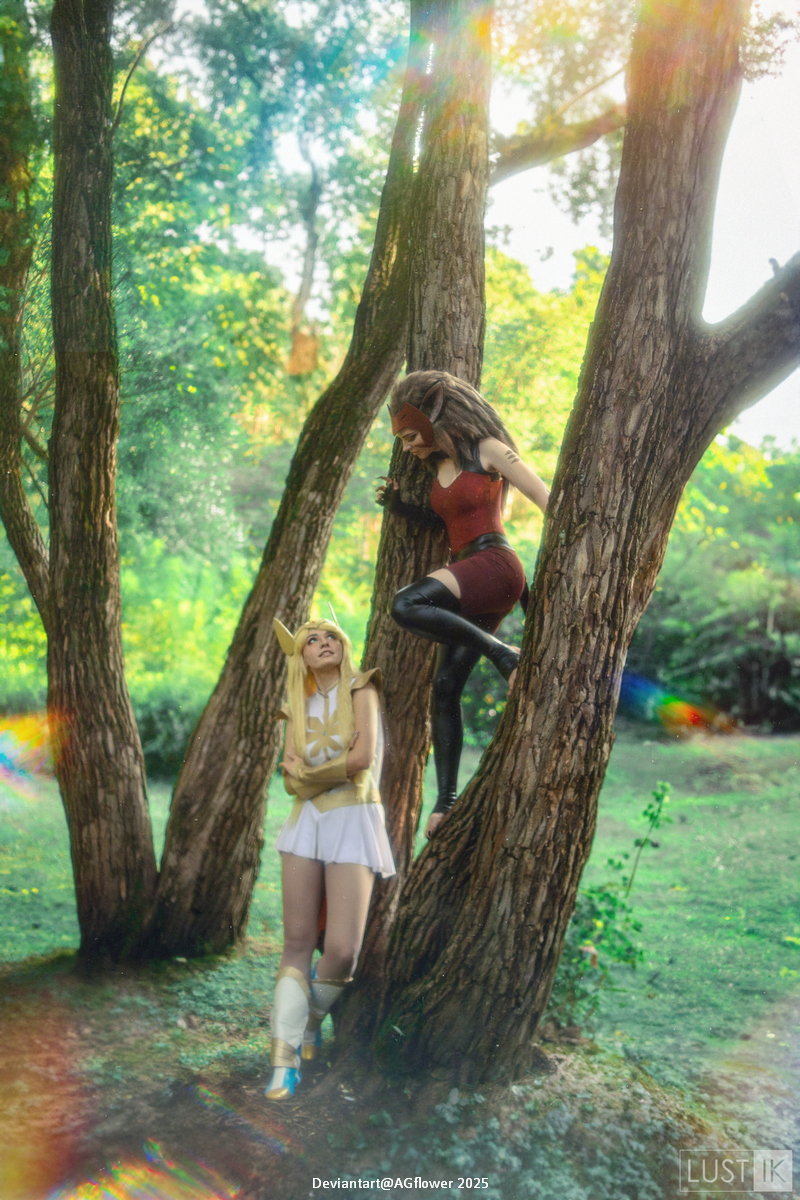 Two individuals in cosplay, one in a light-colored outfit with a hood and the other in a dark outfit, poses on and around trees in a lush green setting.