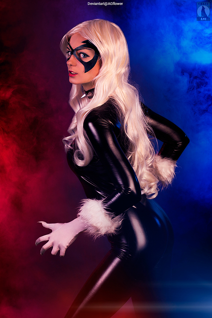 A woman dressed as a comic book character with long blonde hair, wearing a black costume and mask, posing dramatically against a colorful smoke background.