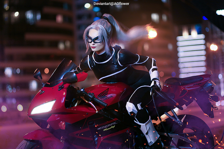 A person in a stylish black bodysuit and mask sits on a red motorcycle, surrounded by a cityscape at night with blurred lights and smoke.