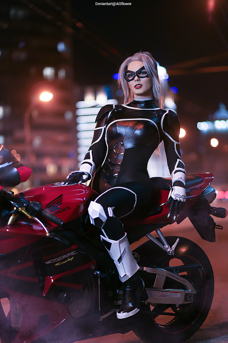 A person dressed in a sleek, black superhero costume with silver accents poses on a red motorcycle at night, illuminated by city lights in the background.