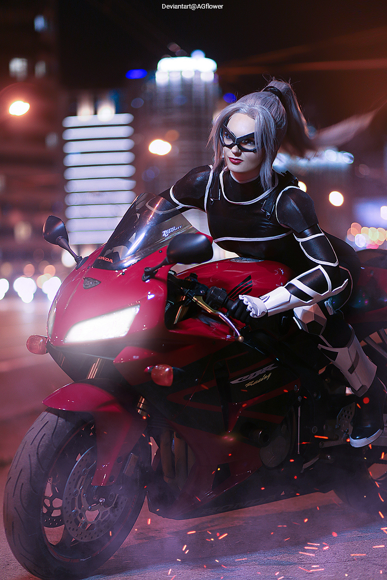 A female character dressed in a black and white costume, wearing sunglasses, poses on a red motorcycle in a bright urban setting at night.