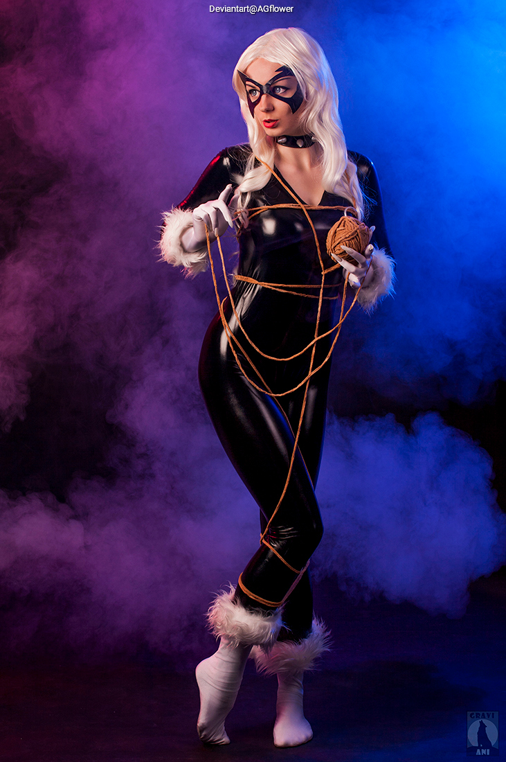 A person in a black catsuit with white fur trim poses dramatically with a ball of yarn in a stylized purple and blue fog background.