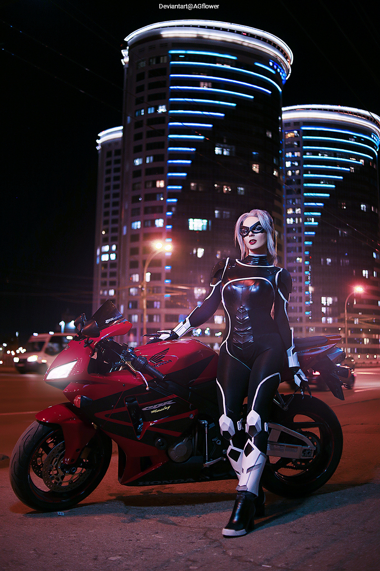 A person in a black and white bodysuit poses on a red motorcycle at night, with modern illuminated buildings in the background.