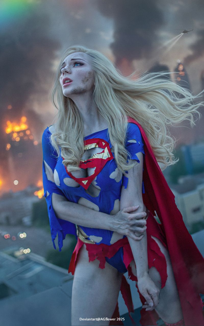 A female superhero character dressed in a torn costume, featuring a blue top with a yellow emblem and a red cape. She stands on a rooftop, looking upwards, with a dramatic sky and urban destruction in the background.