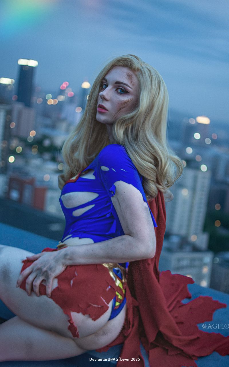 A woman with long blonde hair poses on a rooftop wearing a ripped superhero costume, with a city skyline in the background and a rainbow in the sky.
