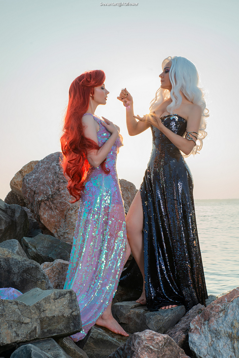 Two cosplayers on rocky beach during sunset, one wearing a shimmering iridescent gown and the other in a black sequined dress.