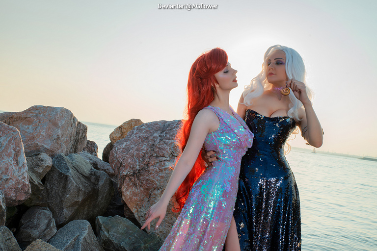 Two women in glamorous costumes pose by the water, with one wearing a sparkling pink dress and the other in a black sequined gown, surrounded by rocks and a serene sea background.