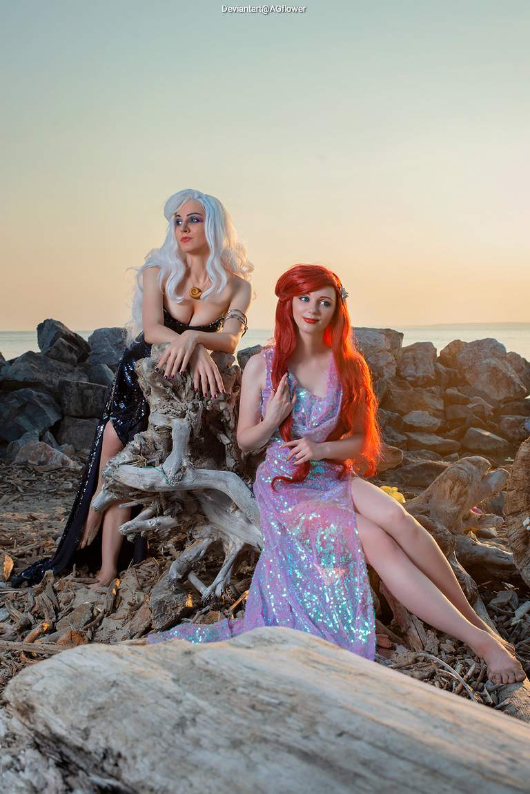 Two women in elaborate costumes sitting on rocks by the shore during sunset. One has long silver hair and a dark dress, while the other has long red hair and a shimmering pink dress.