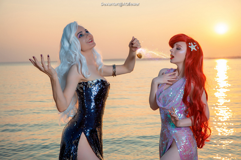 Two women in glamorous dresses pose by the water during sunset. One has long white hair and is holding a seashell, while the other has long red hair and is wearing a sparkling purple dress.