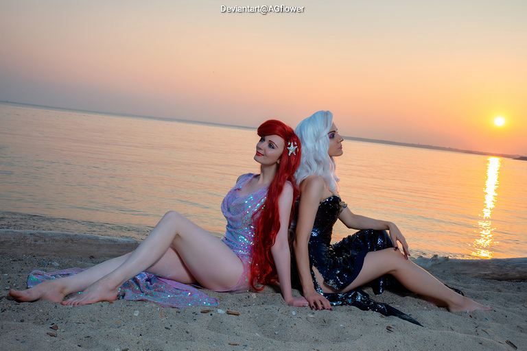 Two women in glamorous dresses sit on a beach at sunset, one with long red hair and the other with silver hair, both looking serene.